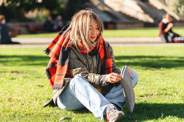 Woman sitting on grass in park, wearing a scarf and enjoying a sunny day while adjusting her hat