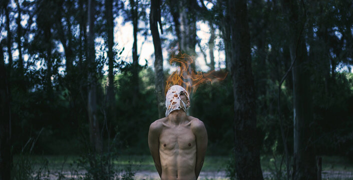 Portrait of a shirtless man standing in a forest wearing a burning mask
