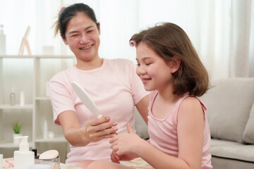 Asian mother teaching Caucasian daughter how to apply makeup in front of mirror. Smiling, sharing joyful beauty lesson at home. Cozy setting with natural light, warm family bonding moment.