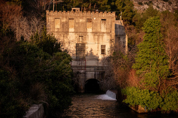 An old, abandoned building with a castle-like facade stands by a flowing river, surrounded by lush...
