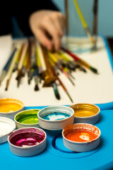 A hand selects a paintbrush from a colorful art setup with vibrant paints on a blue tray. Ideal for showcasing creativity, painting supplies, and artistic projects in action.