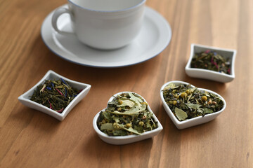 White tea cup with saucer and herbal teas in bowls on wooden background