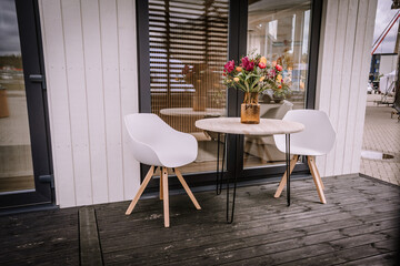 Two wooden chairs and a matching table are placed on the terrace of a charming white wooden house. The table is adorned with a vase filled with colorful flowers, adding a vibrant touch to the scene