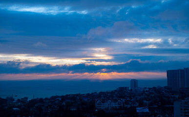 Scenic coastal city skyline at sunrise with dramatic clouds over the ocean. Sun rays breaking through clouds above urban buildings and houses. Morning sky and sea view.