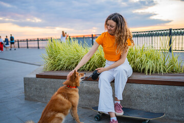 Young woman with curly hair, wearing an orange t-shirt and white pants, sits on her skateboard by a...