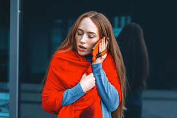 Woman in red shawl talking on phone while standing outside modern building in cool weather during...