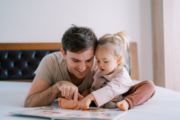 Dad and little girl are reading a book of fairy tales on the bed, running their fingers through the pages