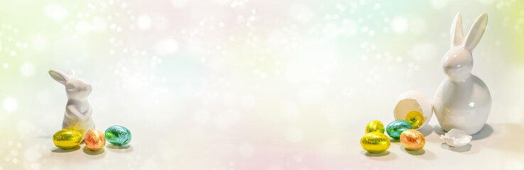 Two white ceramic Easter bunnies left and right with small colorful chocolate Easter eggs against a pastel colored light background, wide panoramic format, copy space