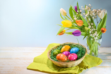 Colorful Easter eggs in a basket and a spring bouquet with tulips on light wood against a blue...