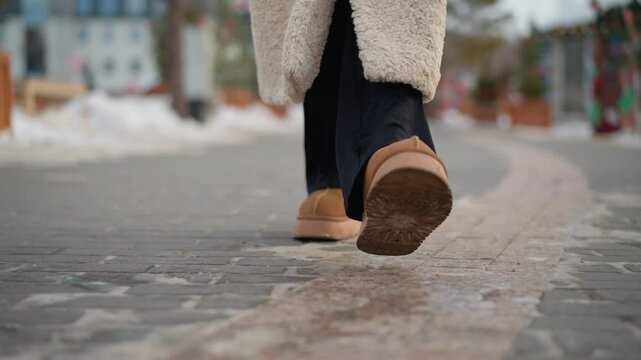 Close-up view of girl wearing warm beige winter coat and brown suede boots walking on snowy path, textured sole of boots visible as she steps forward on cold pavement with snow-covered surroundings