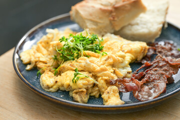 Scrambled eggs with cress garnish, crispy roasted bacon and bread and on a plate on a wooden table, hearty breakfast, lunch or dinner, selected focus