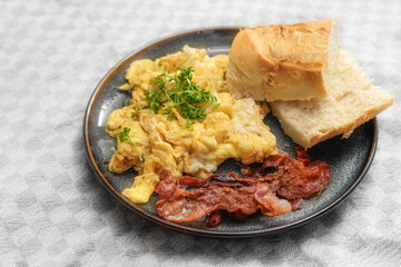 Scrambled eggs with crispy bacon, bread and cress garnish on a plate and a light gray tablecloth,...