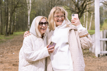 Fototapeta premium Two cheerful senior caucasian women taking a selfie together in a park, smiling and enjoying the moment. They are dressed in cozy, neutral-colored outfits with hoodies and sunglasses