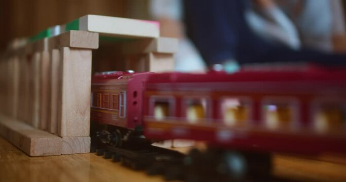 Electric Train for Children &ndash; toy emerges from a tunnel made of wooden blocks, with a blurred silhouette of a playing child in the background. | 4K