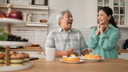 Smiling grandfather with casual cloth sitting at table while grandmother presenting breakfast. Senior couple talking and spending time or supporting together. Healthy relationship concept. Myrmidon.