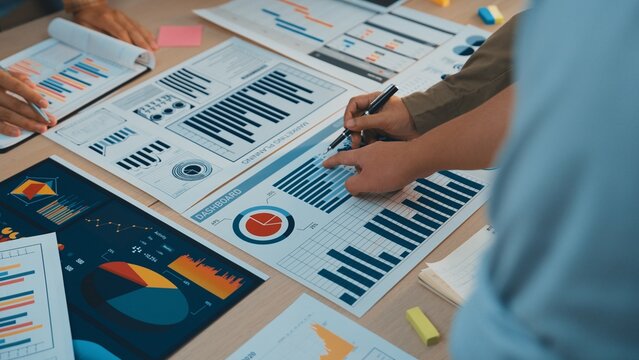 A group of professionals collaborates over various business charts and graphs, engaging in lively discussion and strategic planning for future projects and performance evaluation. SACTR