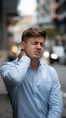 Fototapeta premium Young man in light-blue shirt experiencing neck pain outdoors, showcasing discomfort and health issues, representing stress or physical strain