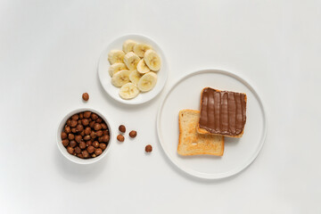 Plate of bread and chocolate spread, hazelnuts and banana slices on a light background. A popular breakfast or snack. View from above.