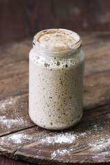 Sourdough bread yeast in a jar with wheat flour and bread loaf on dark wooden rustic background, fermentation, natural homemade baking and cuisine concept