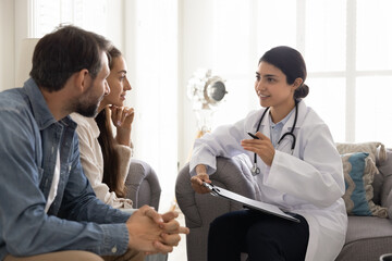 Positive young Indian doctor woman visiting couple at home, writing medical records, filling...