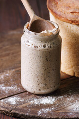 Sourdough bread yeast in a jar with wheat flour and bread loaf on dark wooden rustic background, fermentation, natural homemade baking and cuisine concept