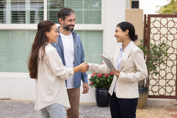 Happy couple of customers and young realtor woman shaking hands outside of house. New homeowners...