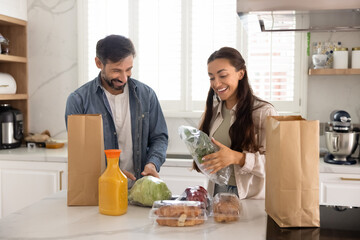 Cheerful married couple in love unpacking bags with food from grocery store, talking, laughing,...