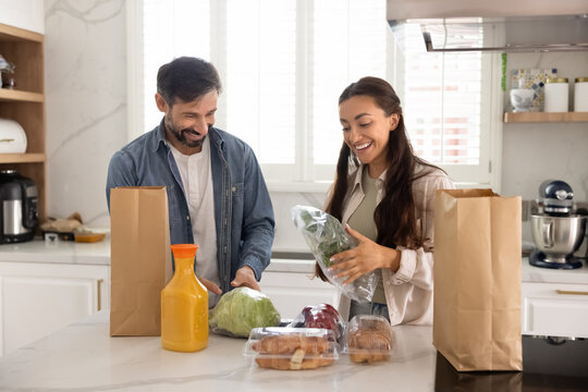 Cheerful married couple in love unpacking bags with food from grocery store, talking, laughing, enjoying leisure, weekend at home, domestic activity, producing fresh vegetables from paper packets
