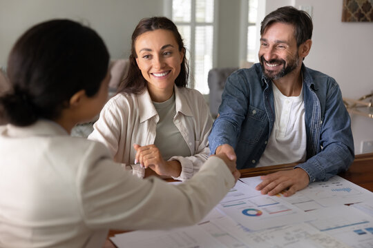 Happy couple of clients thanking financial adviser for consultation, shaking hands over paper analytics on table, giving handshake to personal investment planner, smiling, finishing meeting