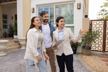 Indian realtor woman showing house and yard for buying to happy couple, holding tablet, pointing...