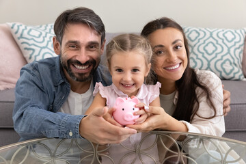 Happy mom and dad hugging little daughter kid holding toy ceramic piggybank, looking at camera, smiling, posing for portrait, making investment, saving money for kids education
