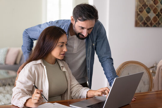 Positive husband and wife using online bank application on laptop for payment domestic bills, taxes, budget management, sitting and standing at computer, pointing and looking at screen - Powered by Adobe