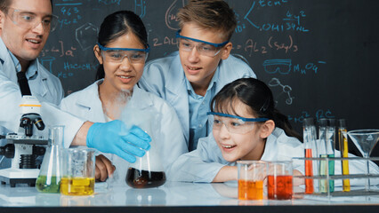 Girl looking at beaker while doing experiment at blackboard with chemistry theory in STEM class. teacher and group of highschool student with mixed races doing science activity together. Edification.