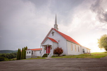 Sun breaking through the clouds behind a church on the top of a hill with a red roof.