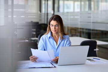 Businesswoman of middle age manager executive reading paper using laptop computer at work. Busy...