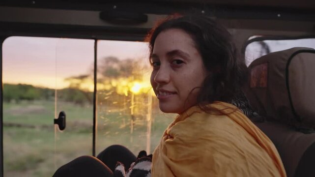 Young woman looking at camera as sun sets behind her during a ride in safari car. Female tourist smiles having fun in perfect vacation in Africa as vehicle drives on dirt road during sunset or sunrise