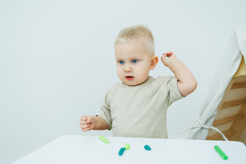 A little one-year-old boy is sculpting plasticine while standing at the table. The child is playing...