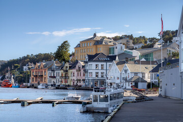 Fototapeta premium Quaint houses line the waterfront, reflecting vibrant colors against a clear blue sky. Boats rest peacefully in the harbor, creating a serene atmosphere in Farsund, picturesque Norwegian coastal town