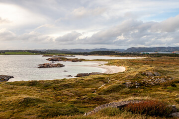A tranquil coastal landscape showcases gentle sandy shores and rugged rocky formations in Farsund, Norway. The sky is filled with soft clouds, enhancing the serene atmosphere of this picturesque bay.