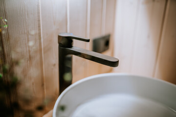 A close-up of a modern black faucet and white ceramic sink against a wooden wall in a stylish bathroom interior with soft natural lighting..