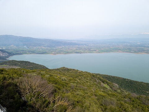 Panoramic view of a vast lake surrounded by green hills and distant mountains under a hazy sky.