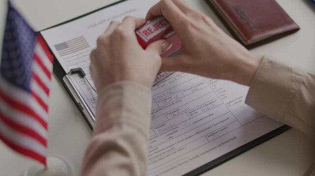 Hands of female consular officer stamping rejected mark on visa application form after reviewing it at desk with American flag and passport nearby. Close-up shot