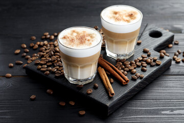 Tasty latte macchiato in glasses, coffee beans and cinnamon on black wooden table, closeup
