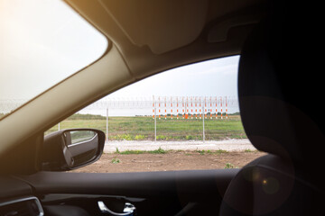 Sunlit view from a car showing a fenced airport area with orange and white runway markers. Ideal for capturing the essence of travel, security, and aviation journeys.