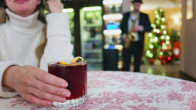 Close up of a woman holding a negroni cocktail on a red and white tablecloth at a restaurant with a Christmas tree on the background