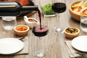 Pouring red wine into glass at wooden table with tasty food, closeup