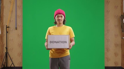 Man holding a charity donation box with a happy expression on green background. Encouraging people to support humanitarian initiatives. Greenscreen, awareness, fundraiser, relief, kindness