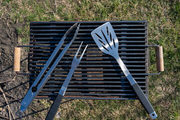 Set of BBQ tools. Barbecue tools (tongs, carving fork, spatula). Close-up of an open empty barbecue grill with a large fork, spatula and tongs on top outdoors.