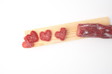 Heart-shaped slices of cured sausage on wooden board with whole sausage. Minimalistic food presentation, catering, social media content. Valentines Day food concept. Flat lay with copy space