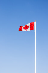 The image shows a Canadian flag fluttering in the wind. The red and white colors of the flag stand out vibrantly under the light of day, while the wind sweeps through it, creating a fluid movement.